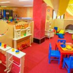 Ein farbenfrohes Indoor-Spielzimmer mit Kindertischen, Stühlen, Spielzeug, Spielküchen, weichen Blöcken und einem Kletterbereich auf rotem Teppichboden.