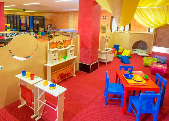 Ein farbenfrohes Indoor-Spielzimmer mit Kindertischen, Stühlen, Spielzeug, Spielküchen, weichen Blöcken und einem Kletterbereich auf rotem Teppichboden.