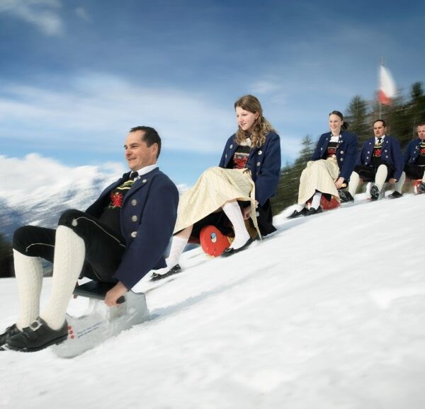 Fünf Menschen in traditioneller Kleidung fahren mit Schlitten einen verschneiten Hügel hinunter, im Hintergrund sind Bäume und Berge unter einem blauen Himmel zu sehen.