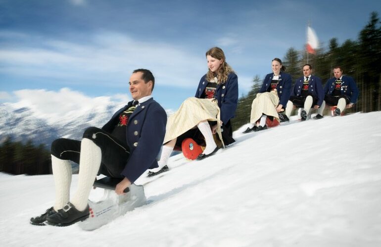 Fünf Menschen in traditioneller Kleidung fahren mit Schlitten einen verschneiten Hügel hinunter, im Hintergrund sind Bäume und Berge unter einem blauen Himmel zu sehen.