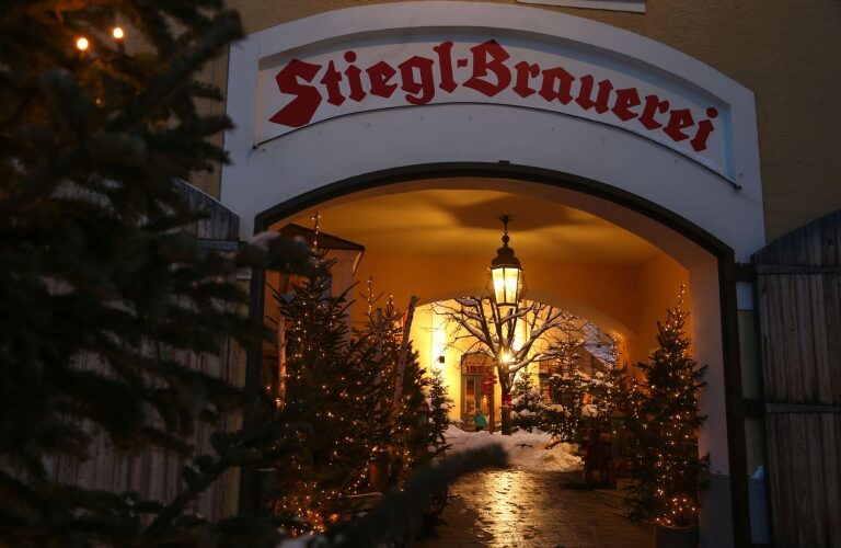 Der Eingang zur Stiegl-Brauerei ist mit Weihnachtsbäumen und Lichtern geschmückt, im Torbogen hängt eine Laterne, im Hintergrund ist Schnee zu sehen.