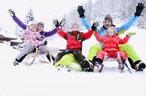 Zwei Erwachsene und drei Kinder in Winterkleidung sitzen auf Schlitten im Schnee, heben die Arme und lächeln, im Hintergrund sind schneebedeckte Bäume zu sehen.