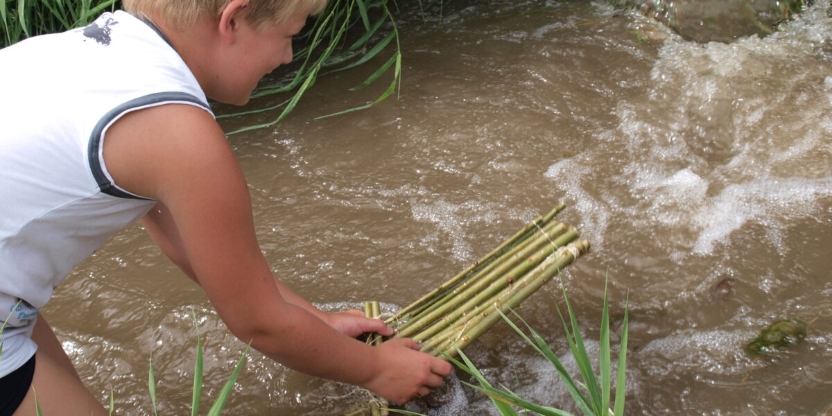 Ein Kind kniet an einem schlammigen Bach und lässt ein kleines selbstgebautes Floß aus Stöcken ins Wasser, umgeben von grünem Gras.