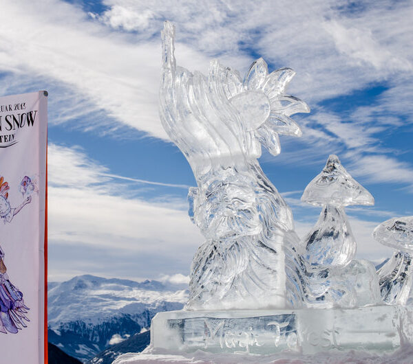 Eine klare Eisskulptur einer bärtigen Figur mit einem hohen Hut und zwei Pilzen wird im Freien auf Schnee ausgestellt, mit Bergen und einem „Art on Snow“-Veranstaltungsbanner im Hintergrund.