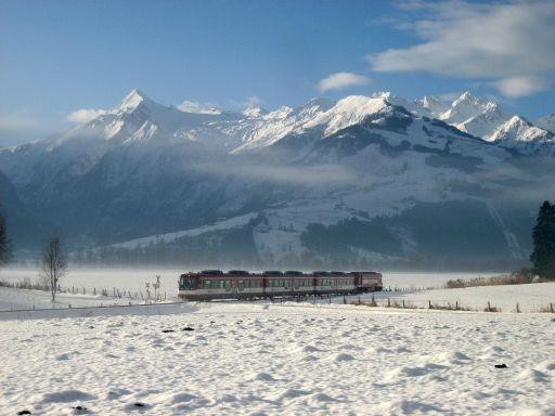 Ein roter Zug fährt durch eine verschneite Landschaft mit hohen, schneebedeckten Bergen im Hintergrund unter einem teilweise bewölkten Himmel.