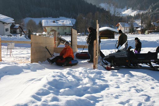 Mehrere Menschen in Winterkleidung sitzen auf Snowtubes und bereiten sich darauf vor, von einem Schneemobil über ein verschneites Feld in der Nähe von Häusern und Bäumen gezogen zu werden.