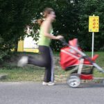 Eine Frau schiebt in Bewegung einen roten Kinderwagen eine Straße entlang, im Hintergrund sind Grünflächen und ein gelbes Hinweisschild zu sehen.