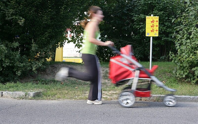 Eine Frau schiebt in Bewegung einen roten Kinderwagen eine Straße entlang, im Hintergrund sind Grünflächen und ein gelbes Hinweisschild zu sehen.