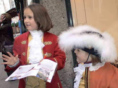 Zwei Kinder in historischen Kostümen stehen auf einer Straße. Das eine hält Flugblätter und spricht, das andere trägt einen Dreispitz mit weißen Federn.