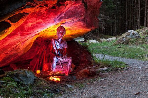 Eine rot und weiß bemalte Skulptur in Menschenform steht unter einem Felsvorsprung, der von roten und gelben Lichtern beleuchtet wird, neben einem Waldweg.