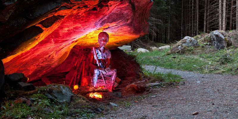 Eine rot und weiß bemalte Skulptur in Menschenform steht unter einem Felsvorsprung, der von roten und gelben Lichtern beleuchtet wird, neben einem Waldweg.