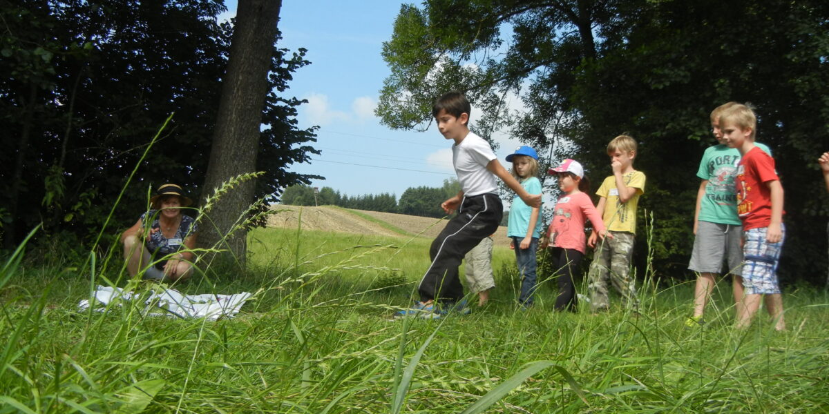 Mehrere Kinder stehen auf einer Wiese im Freien, während ein Junge nach vorne springt. Ein Erwachsener sitzt in der Nähe auf dem Boden und beobachtet. Im Hintergrund sind Bäume und Felder zu sehen.