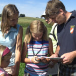 Vier Personen und ein Kind stehen an einem sonnigen Tag draußen auf einer Wiese und schauen auf eine Karte und ein GPS-Gerät.
