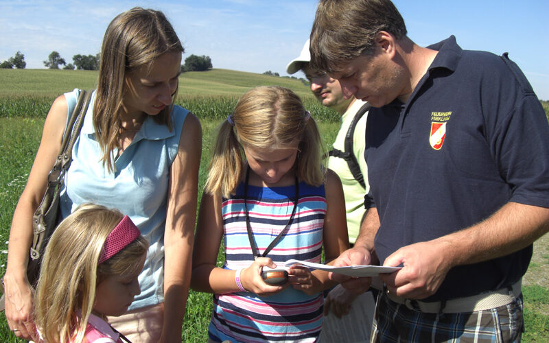 Vier Personen und ein Kind stehen an einem sonnigen Tag draußen auf einer Wiese und schauen auf eine Karte und ein GPS-Gerät.