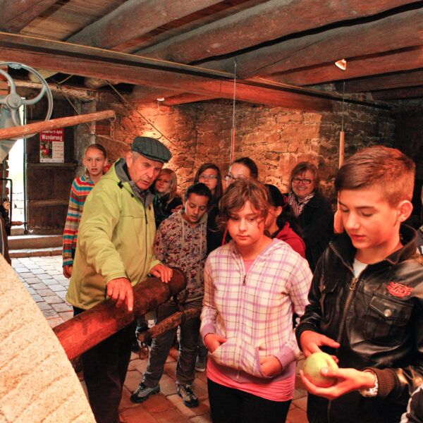 Eine Gruppe von Kindern und ein Erwachsener stehen in einem rustikalen Gebäude mit Backsteinwänden und Holzbalken und betrachten alte Maschinen.