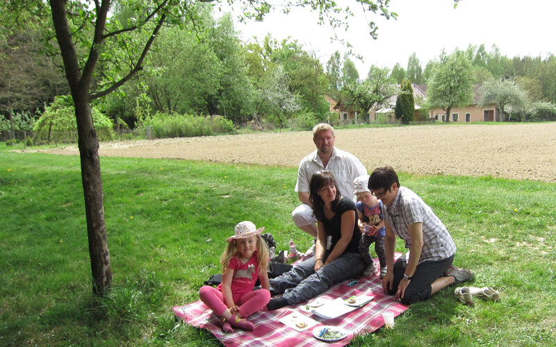Eine fünfköpfige Familie sitzt auf einer Decke und macht ein Picknick unter einem Baum in der Nähe eines Feldes. Auf der Decke stehen Teller mit Essen, im Hintergrund sind Häuser zu sehen.