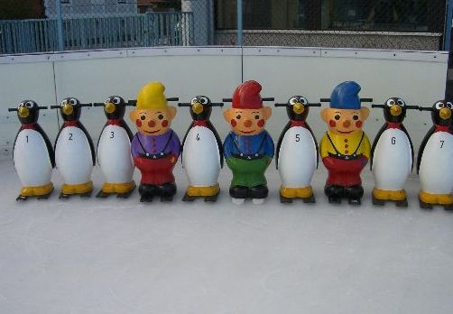 Auf einer Eisbahn sind sieben Pinguin-Laufhilfen, jeweils mit einer Nummer versehen, und drei bunte Clown-Laufhilfen aufgereiht.