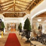 Überdachter Eingang mit Holzbalken, rotem Teppich, Topiary-Pflanzen, schwarzen, verzierten Bänken und Glastüren, die in einen hellen Innenraum führen.