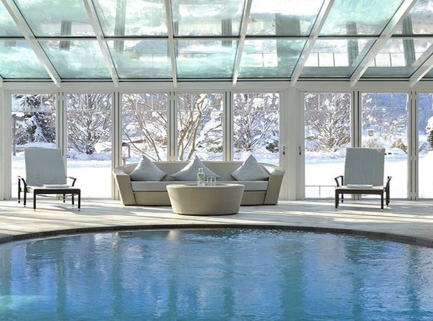 Sitzbereich im Innenbereich mit rundem Tisch und Stühlen neben dem Pool, umgeben von Glaswänden, die eine verschneite Landschaft nach draußen zeigen.