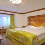 Hotelzimmer mit einem großen Doppelbett, Holzmöbeln, einem an der Wand montierten Fernseher, zwei grünen Sesseln am Fenster und neutraler Einrichtung mit grünen Akzenten.