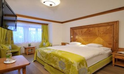 Hotelzimmer mit einem großen Doppelbett, Holzmöbeln, einem an der Wand montierten Fernseher, zwei grünen Sesseln am Fenster und neutraler Einrichtung mit grünen Akzenten.