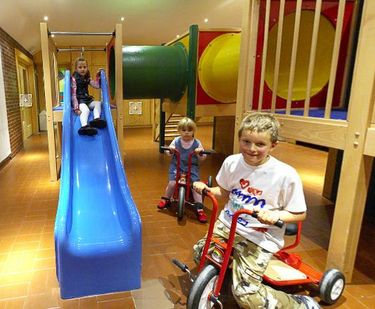 Drei Kinder spielen drinnen; eines rutscht eine blaue Rutsche hinunter, und zwei andere fahren mit Dreirädern in der Nähe eines hölzernen Spielgerüsts mit einem Tunnel.