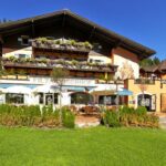 Ein Hotel im Chaletstil mit Balkonen, Blumenkästen und Sitzgelegenheiten im Freien ist umgeben von grünem Gras und Bäumen unter einem klaren Himmel.