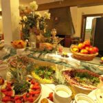 Ein Buffet mit verschiedenen Obstsorten, Salaten, Aufschnitt und Desserts, dekorativen Blumen und einem Korb mit Äpfeln steht in einem gut beleuchteten Essbereich.