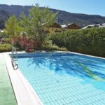 Außenschwimmbad mit klarem, blauem Wasser, umgeben von grünen Hecken, Bäumen und Bergen im Hintergrund unter einem sonnigen Himmel.