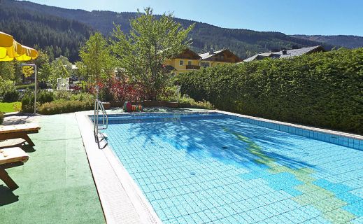 Außenschwimmbad mit klarem, blauem Wasser, umgeben von grünen Hecken, Bäumen und Bergen im Hintergrund unter einem sonnigen Himmel.