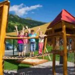 Vier Kinder stehen auf einer hölzernen Brücke auf einem Spielplatz im Freien und heben die Arme. Im Hintergrund sind ein Gebäude und Hügel unter blauem Himmel zu sehen.