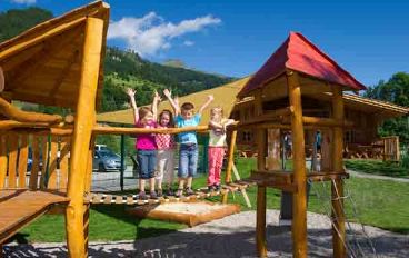 Vier Kinder stehen auf einer hölzernen Brücke auf einem Spielplatz im Freien und heben die Arme. Im Hintergrund sind ein Gebäude und Hügel unter blauem Himmel zu sehen.