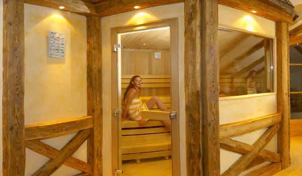 Eine Frau mit einem Handtuch sitzt in einer Holzsauna mit Glasfenstern und rustikaler Holzverkleidung.