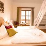 Ein Hotelzimmer mit einem großen Bett, Kissen, Bademänteln, einem an der Wand montierten Fernseher, einem Schreibtisch mit Stühlen, einer Treppe und einem Fenster mit Vorhängen, die das Tageslicht hereinlassen.