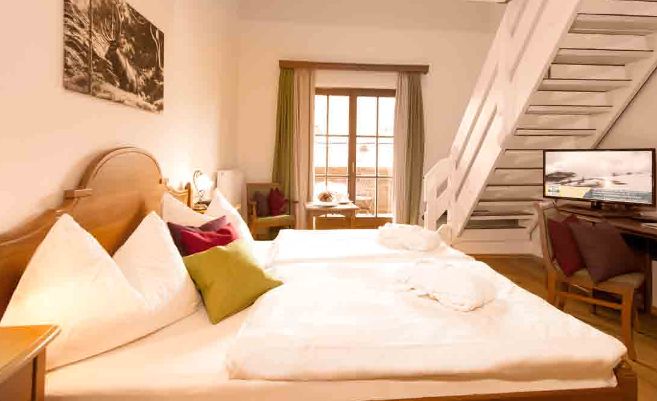 Ein Hotelzimmer mit einem großen Bett, Kissen, Bademänteln, einem an der Wand montierten Fernseher, einem Schreibtisch mit Stühlen, einer Treppe und einem Fenster mit Vorhängen, die das Tageslicht hereinlassen.