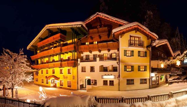 Ein warm beleuchtetes Hotel im alpinen Stil ist nachts von Schnee umgeben, mit Balkonen, Holzbalken und verschneiten Bäumen im Vordergrund.