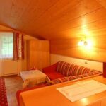 Gemütliches Zimmer mit holzgetäfelter Decke, rot gestreiftem Sofa, kleinem Tisch, Kleiderschrank und großem Fenster mit Vorhängen, die Tageslicht hereinlassen.