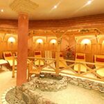 Ein runder Spa-Entspannungsraum mit hölzernen Liegestühlen, einer dekorativen Säule in der Mitte und einem mit Steinen gesäumten Trockenbrunnen. Warme Beleuchtung und Blumendekoration sind vorhanden.