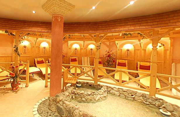 Ein runder Spa-Entspannungsraum mit hölzernen Liegestühlen, einer dekorativen Säule in der Mitte und einem mit Steinen gesäumten Trockenbrunnen. Warme Beleuchtung und Blumendekoration sind vorhanden.