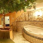 Wellnessbereich im Innenbereich mit einem großen Whirlpool aus Stein, Mosaikfliesenwänden, einem Baum in der Mitte mit einer runden Bank und warmer Beleuchtung.