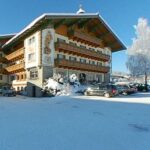 Ein traditionelles alpenländisches Gebäude mit Balkonen und Wandmalereien, umgeben von Schnee und geparkten Autos, unter einem klaren blauen Himmel.