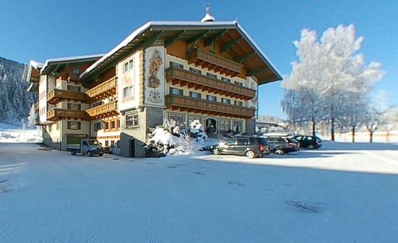 Ein traditionelles alpenländisches Gebäude mit Balkonen und Wandmalereien, umgeben von Schnee und geparkten Autos, unter einem klaren blauen Himmel.