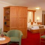 Hotelzimmer mit einem Doppelbett, Holzmöbeln, einem Tisch mit zwei Stühlen, einem Sessel und einer Flasche Wein mit Gläsern auf einem Tisch. Roter Teppich und neutrale Wände.