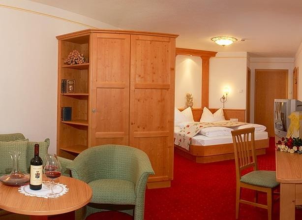 Hotelzimmer mit einem Doppelbett, Holzmöbeln, einem Tisch mit zwei Stühlen, einem Sessel und einer Flasche Wein mit Gläsern auf einem Tisch. Roter Teppich und neutrale Wände.