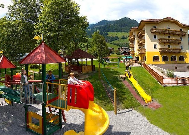Kinder spielen auf bunten Rutschen und Klettergerüsten auf einem Spielplatz neben einem großen gelben Gebäude, mit grasbewachsenen Hügeln und Bergen im Hintergrund.