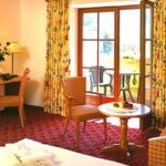 Hotelzimmer mit Bett, Sofa, Tisch mit Obst, Stühlen, Schreibtisch mit Lampe und TV, großem Fenster mit geblümten Vorhängen und einer Tür, die auf einen Balkon führt.