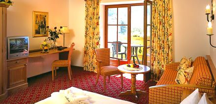 Hotelzimmer mit Bett, Sofa, Tisch mit Obst, Stühlen, Schreibtisch mit Lampe und TV, großem Fenster mit geblümten Vorhängen und einer Tür, die auf einen Balkon führt.