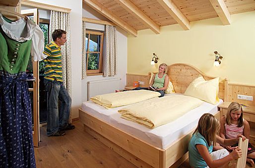 Ein Mann steht an der Tür, während drei Frauen in einem hölzernen Schlafzimmer mit gelber Bettwäsche und natürlichem Licht durch ein Fenster sitzen und sich entspannen.