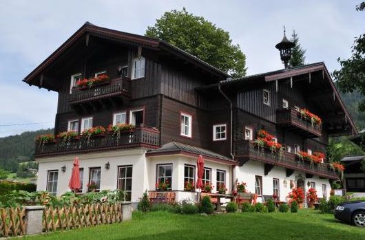 Ein großes Haus im Stil eines dunklen Holzchalets mit Balkonen, die mit Blumenkästen geschmückt sind, und einem Vorgarten in einer grünen, bergigen Gegend.