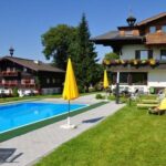 Außenpool mit gelben Sonnenschirmen, Liegestühlen und zwei Gebäuden im alpinen Stil, umgeben von grünem Gras und Bäumen unter einem klaren blauen Himmel.